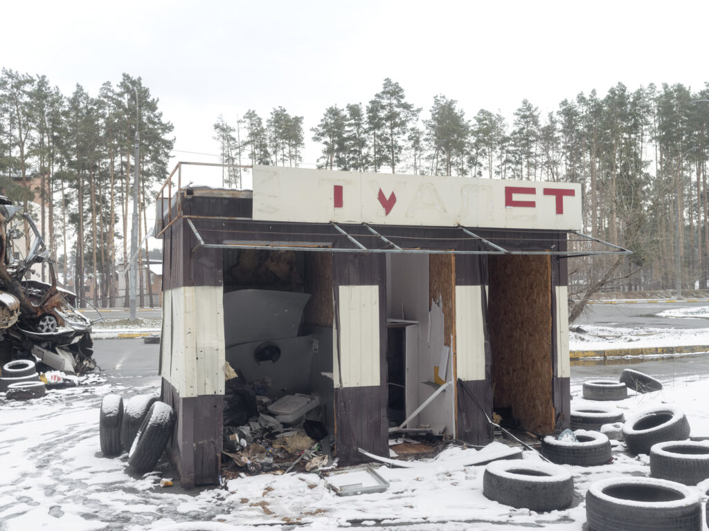 Une petite cabine endommagée, ouverte sur son intérieur, se dresse parmi des pneus et des véhicules détruits dans la neige.