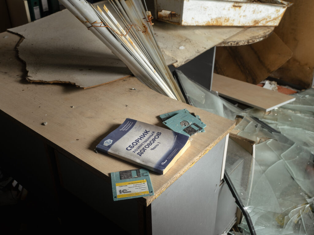 Sur une table poussiéreuse, un manuel et des disquettes abandonnés reposent près de cadres métalliques et de vitres brisées.