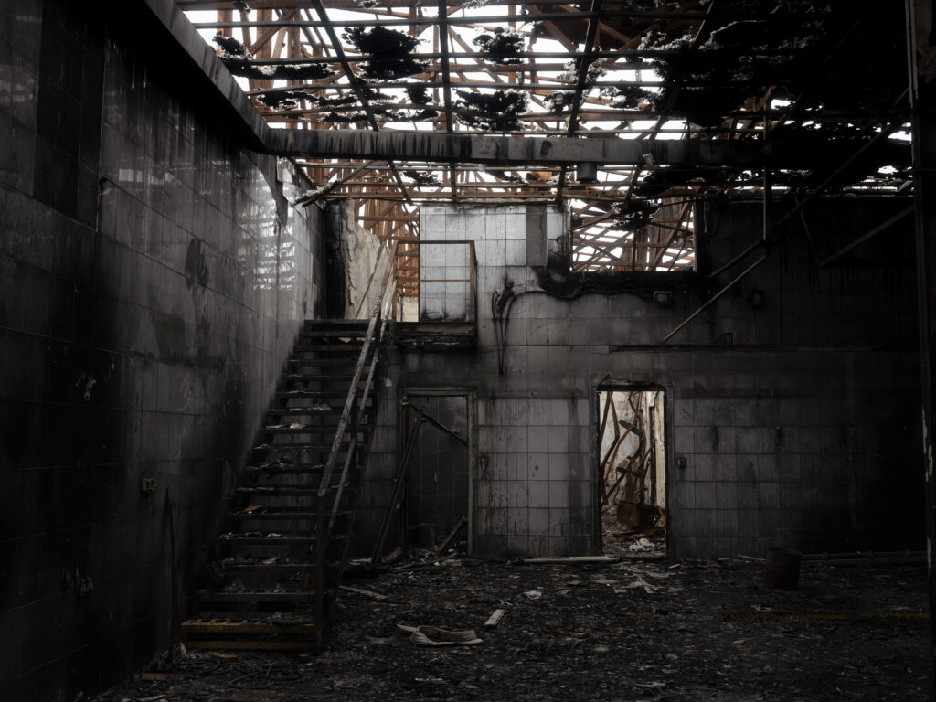 Dans un bâtiment noirci, un escalier métallique monte vers une charpente de toiture percée laissant entrer la lumière.