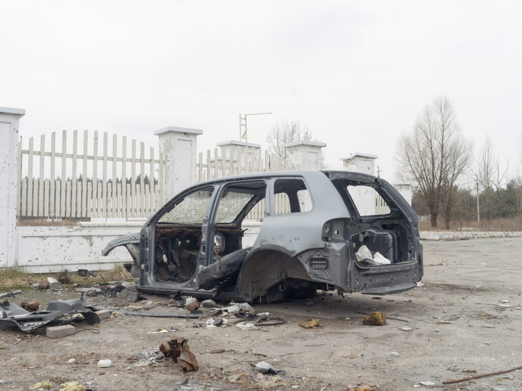 La carcasse noircie d'une voiture sans roues est abandonnée devant une grille blanche, entourée de débris.