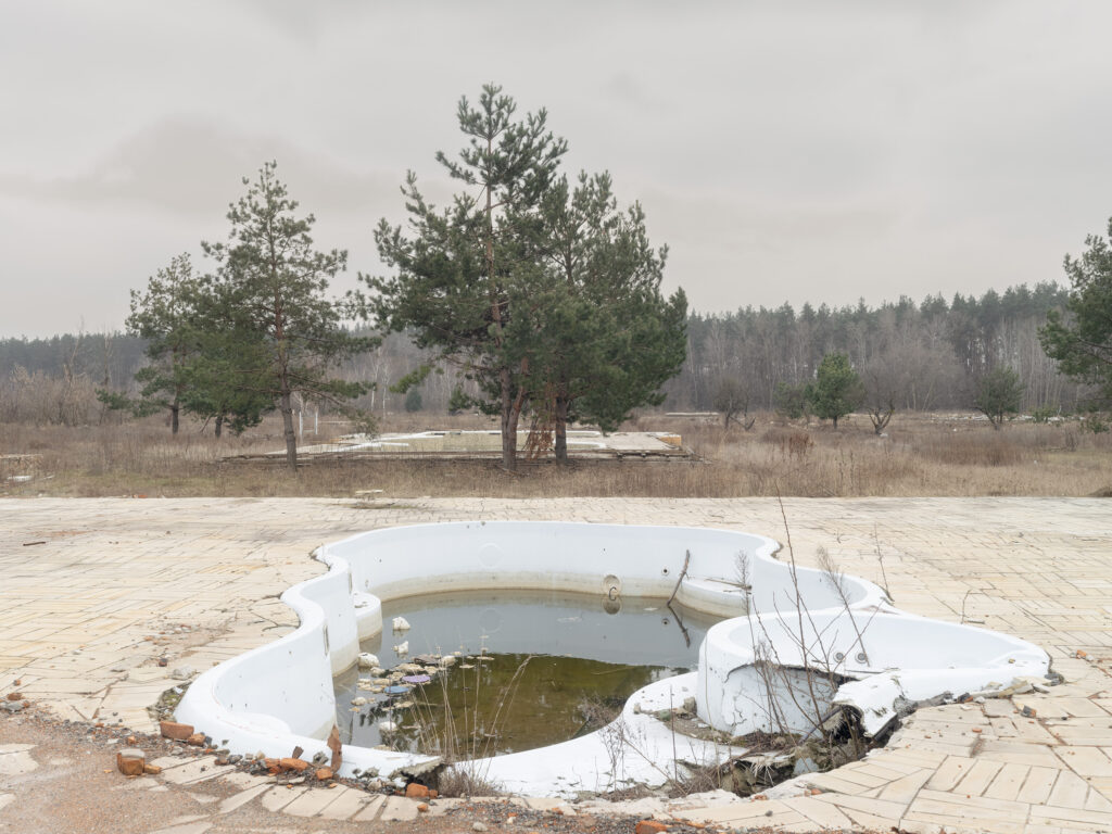 Un bassin circulaire à moitié rempli d'eau, aux rebords brisés, se trouve dans un espace pavé désert près d'une forêt.