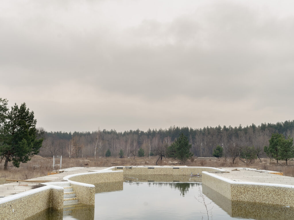Un grand bassin géométrique abandonné, rempli d'eau stagnante, s'étend devant une lisière de pins sous un ciel couvert.