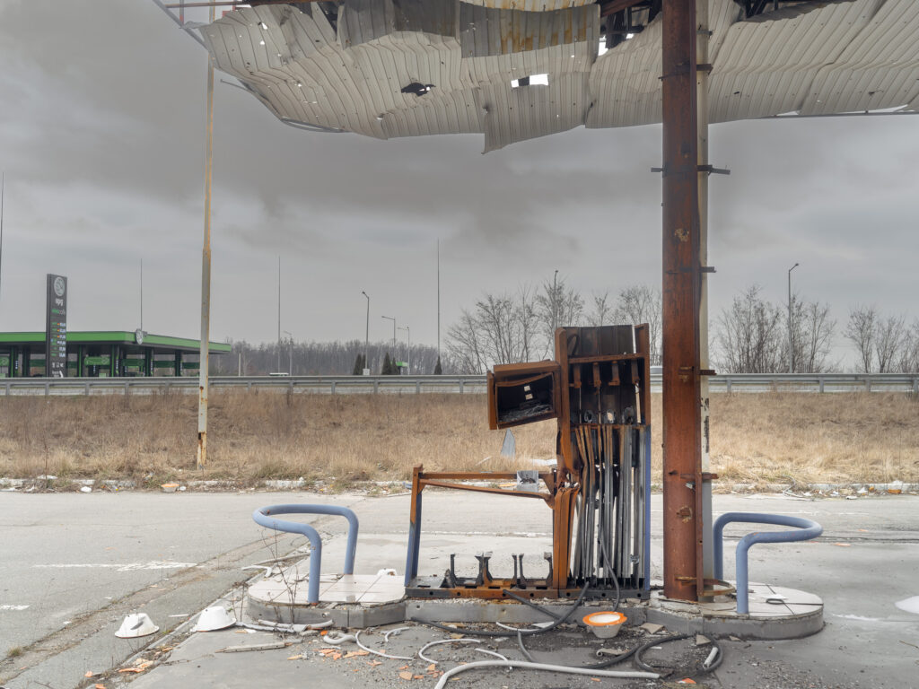 Une ancienne pompe de station-service rouillée se dresse sous un auvent très endommagé, au bord d'une route déserte.