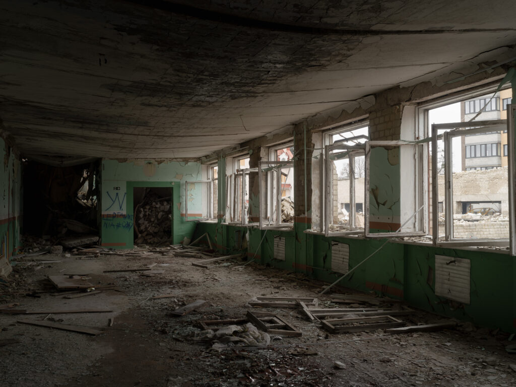 Une salle longue et vide aux murs verts, avec des fenêtres brisées, des radiateurs et des fragments de plafond au sol.