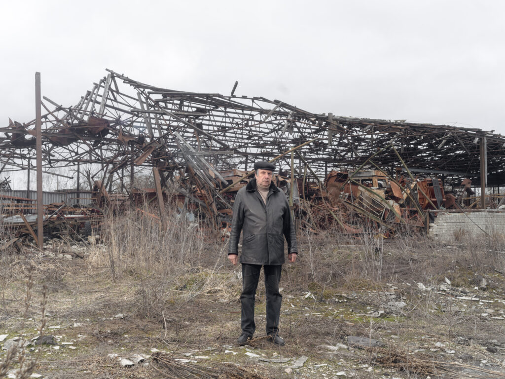 Un homme se tient seul devant l'ossature effondrée d'un vaste bâtiment métallique, au milieu des herbes.