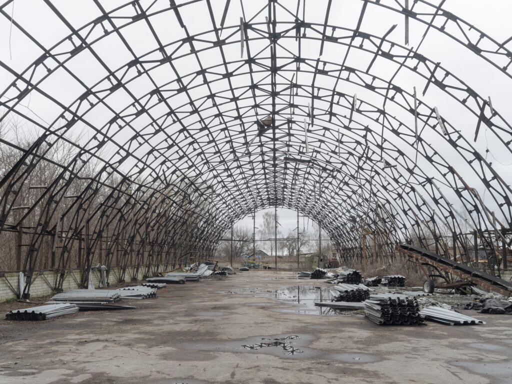 Un immense squelette de serre ou de hangar forme une succession d'arceaux métalliques au-dessus d'un sol abandonné.