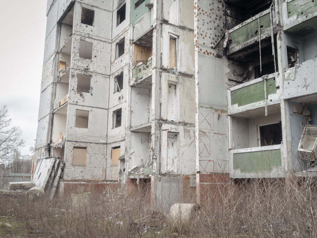 Un immeuble résidentiel aux balcons et aux murs détruits se dresse derrière une bande de végétation sèche.