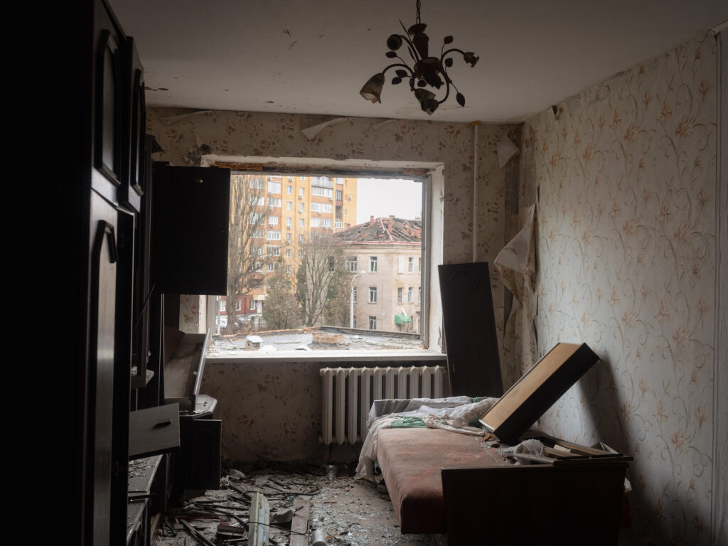 Une chambre endommagée, avec une fenêtre ouverte sur des immeubles, des meubles cassés et des débris sur le sol.