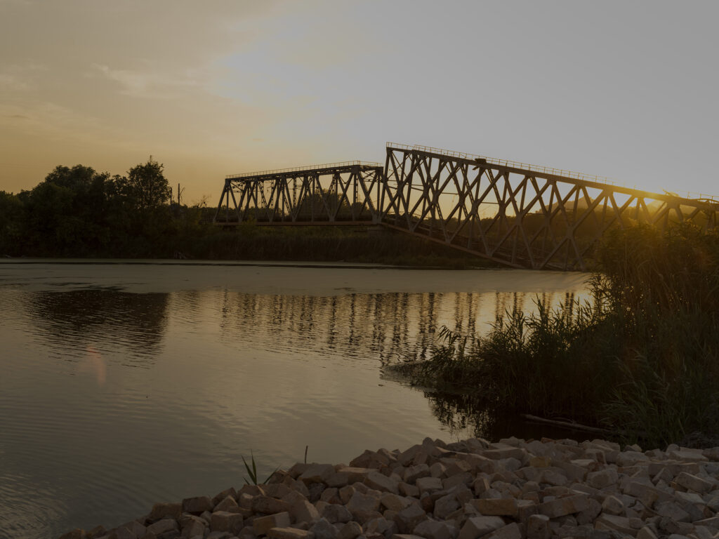 Donbas. Un pont métallique en treillis effondré traverse une rivière au coucher du soleil, dont la lumière se reflète dans l'eau.